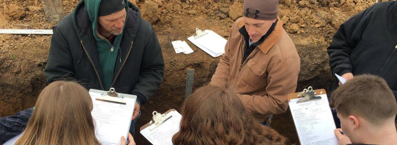 Students taking notes in a soil survey under observation of instructor.