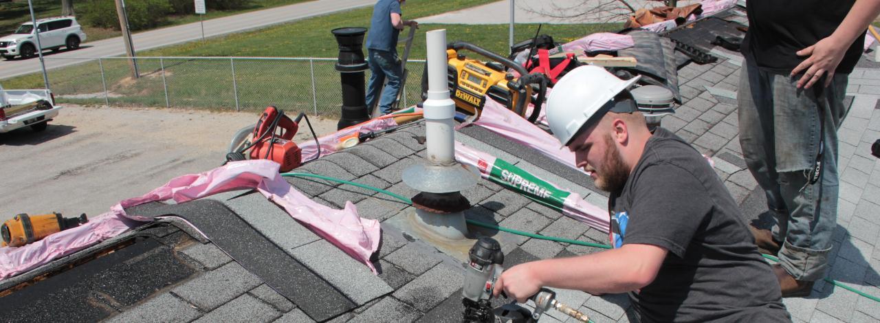 Student on roof operating nail gun to attach shingle to a roof